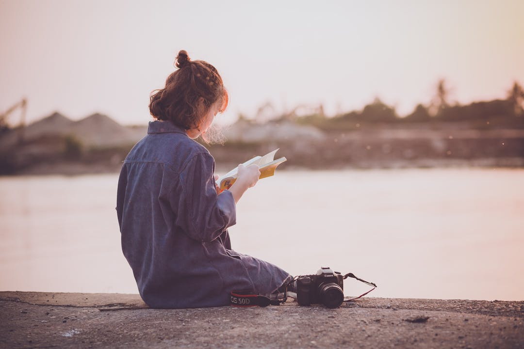 A woman sitting at a still river reading a book
